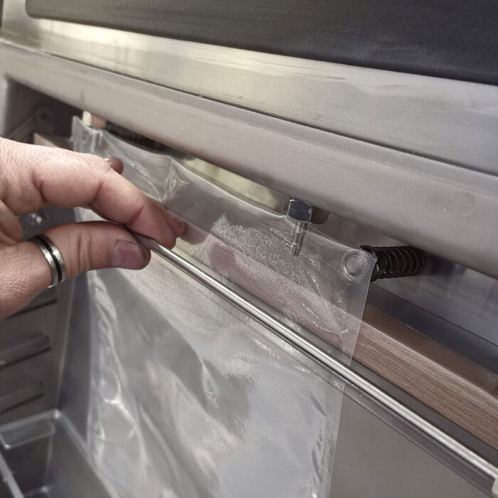 Ręka ze srebrnym pierścieniem na palcu wskazującym umieszcza przezroczystą plastikową torbę na listwie zgrzewającej Komorowej pakowarki próżniowej Eco Vac 420V TB.