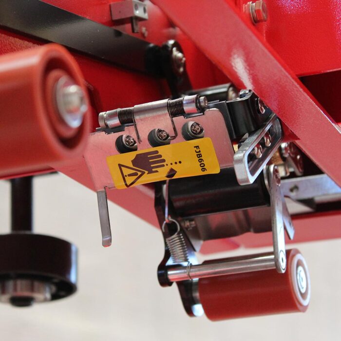Zbliżenie elementu Półautomatyczna zaklejarka Mini Tape 50 SB z czerwoną obudową, metalowymi częściami, rolkami i żółtą etykietą z symbolem dłoni i ostrzeżenia, wskazującym na potencjalne zagrożenie uszczypnięciem.