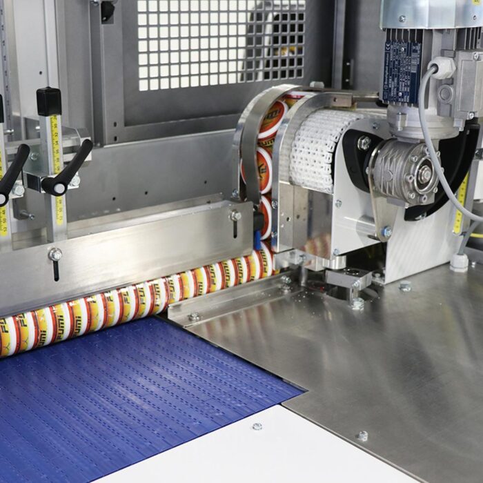 Zbliżenie automatycznej etykieciarki FA800AS i automatycznej zgrzewarki do zgrzewek FA800AS nakładającej okrągłe naklejki na produkty na przenośniku taśmowym, z widocznymi częściami mechanicznymi i etykietami podczas pracy.