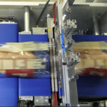 Zbliżenie bochenka chleba pakowanego w folię przez Automat pakujący poziomy FLOWPACK SBM750S-LR, z niebieskimi taśmami przenośnikowymi i widocznymi metalowymi częściami podczas procesu pakowania.