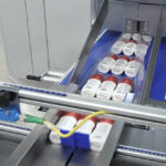 Kilka cylindrycznych pojemników z czerwonymi pokrywkami porusza się na przenośniku taśmowym w Automatycznej pakowarce flow-pack SR750LR, gdzie są one owijane w przezroczystą folię plastikową do wysyłki lub dystrybucji.