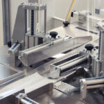 Zbliżenie automatycznej pakowarki flow-pack SR550 wykonanej ze stali nierdzewnej, wyposażonej w linijki, dźwignie i regulowane części do zgrzewania produktów w zakładzie produkcyjnym. Częściowo widoczna przezroczysta folia.