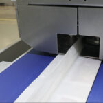 Widok z bliska Automatycznej pakowarki flow-pack SR550LR przetwarzającej i składającej biały papier lub materiał opakowaniowy na przenośniku taśmowym, z widoczną częścią wyposażenia przemysłowego.