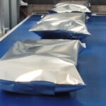 Cztery srebrno-niebieskie próżniowo zgrzewane opakowania są ustawione w rzędzie na niebieskim przenośniku taśmowym, świeżo zapakowane przez Automatyczną pakowarkę flow-pack SR750 w zakładzie pakowania żywności lub produktów.