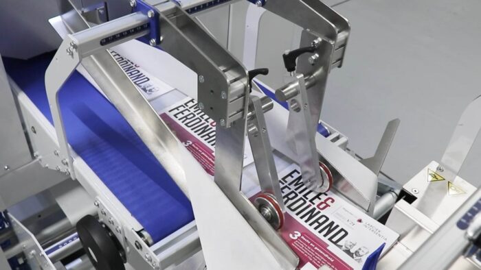 Automat pakujący poziomy FLOWPACK SI550S-LR z metalowymi ramionami i rolkami przetwarza produkty na niebieskim przenośniku taśmowym w fabryce przemysłowej.