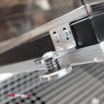 Zbliżenie metalowego zawiasu ze śrubami, sprężyną i przezroczystą pokrywą Zgrzewarki kloszowej do folii Eco Sealer 55. W lewym dolnym rogu znajduje się logo ISG PACK.