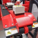 Zbliżenie na Półautomatyczną zaklejarkę Random Tape 50 TBB w kolorze czerwonym, z etykietami ostrzegającymi o ruchomych częściach, czarnym gumowym paskiem i szarym przyciskiem zatrzymania awaryjnego na górze. Etykiety zalecają trzymanie rąk z dala dla bezpieczeństwa.