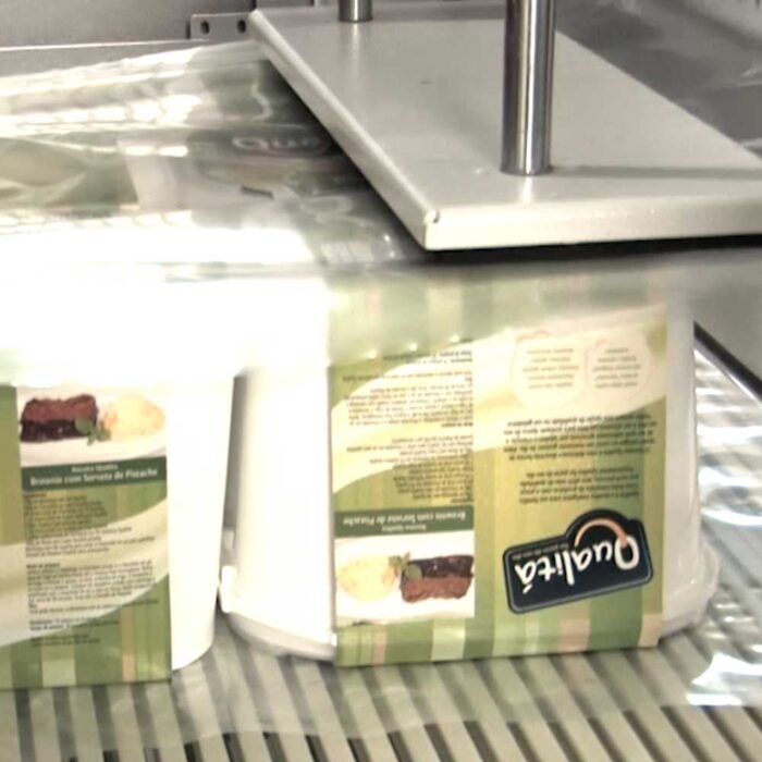 Dwa prostokątne plastikowe pojemniki na żywność z zielonymi i kremowymi etykietami poruszają się wzdłuż przenośnika taśmowego pod maszyną, prawdopodobnie Automatyczną zgrzewarką do zgrzewek FA800AR-350P, w zakładzie pakowania.