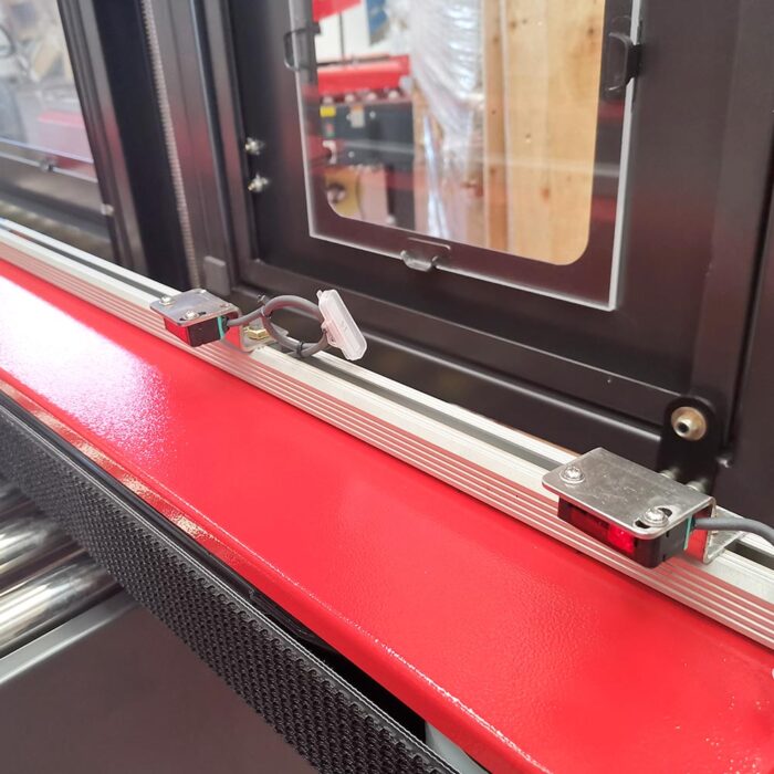 Zbliżenie systemu czujnika liniowego zamontowanego na czerwono-srebrnej szynie z podłączonymi przewodami, obok Półautomatycznej zaklejarki Random Tape 50 SB, w tle panel maszyny z oknem.