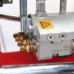 Zbliżenie na Automatyczną zaklejarkę Auto Tape 50 HM, wyposażoną w mosiężne złącza, zwiniętą sprężynę i żółtą naklejkę ostrzegającą o wysokiej temperaturze. Metalowa maszyna jest zamontowana na czerwonej ramie.