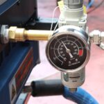 Zbliżenie manometru pokazującego około 80 psi, podłączonego do regulatora z rurami i złączkami na Automatycznej zaklejarce Matic Tape 50 CF HM.