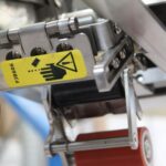 Zbliżenie na półautomatyczną zaklejarkę Eco Tape 50 SB SS z żółtą etykietą ostrzegawczą z ręką, trójkątem ostrzegawczym i gruzem. Widoczne metalowe części i czerwona rolka wskazują na potencjalne zagrożenia podczas pracy.