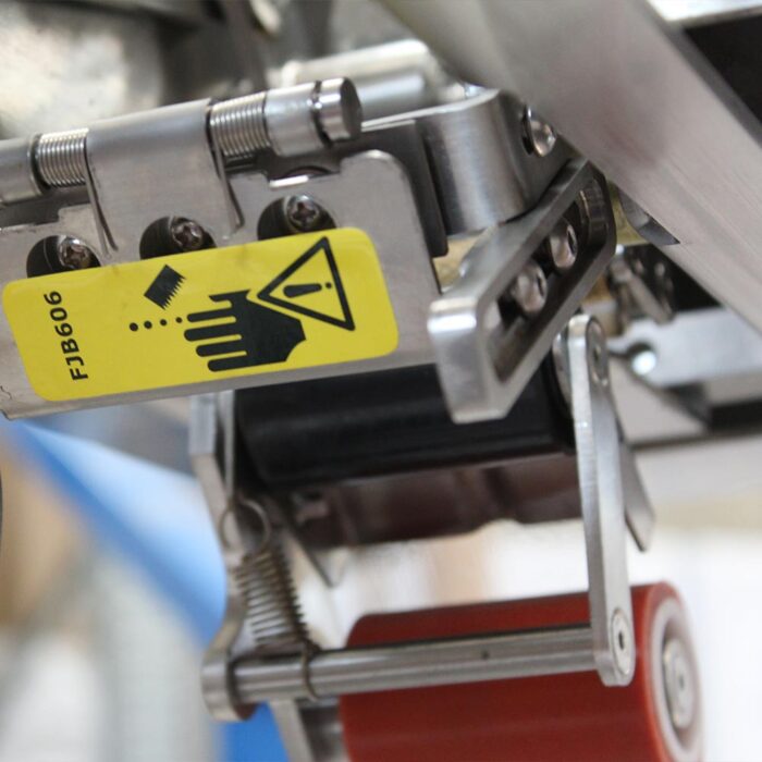 Zbliżenie na półautomatyczną zaklejarkę Eco Tape 50 SB SS z żółtą etykietą ostrzegawczą z ręką, trójkątem ostrzegawczym i gruzem. Widoczne metalowe części i czerwona rolka wskazują na potencjalne zagrożenia podczas pracy.