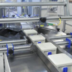 Plastikowe pojemniki z niebieskimi pokrywkami poruszają się wzdłuż przenośnika taśmowego otoczonego metalowymi szynami i maszynami w ramach procesu pakowania Automat pakujący poziomy FLOWPACK SI550S w warunkach przemysłowych.