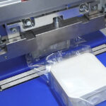 Automat pakujący poziomy FLOWPACK SI750S owija przezroczystą plastikową folię wokół białego prostokątnego pojemnika, gdy ten porusza się wzdłuż niebieskiego przenośnika taśmowego.