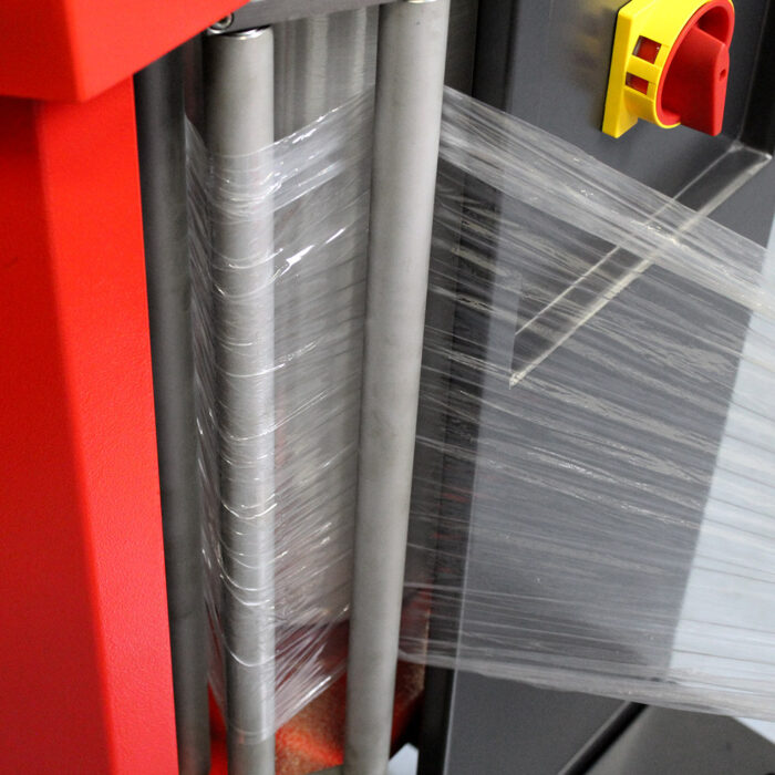 Przezroczysta folia stretch przechodzi między metalowymi rolkami owijarki Owijarka do walizek Baggy Wrap 900 B, która posiada czerwony i żółty przycisk zatrzymania awaryjnego z boku.