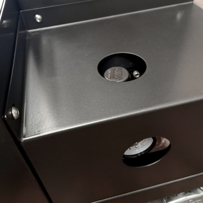 Zbliżenie czarnego metalowego pudełka Box Wrap 800 B Owijarka do paczek, pokazujące okrągłe otwory z monetą i mechanizmem blokującym wewnątrz, widoczne śruby i solidne krawędzie wskazujące na bezpieczną konstrukcję.