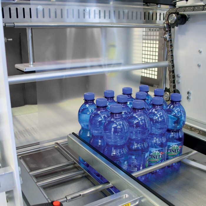 Pakiet niebieskich plastikowych butelek na wodę jest obkurczany na metalowym przenośniku przez Automatyczną zgrzewarkę do zgrzewek FA802AS, zapewniając wydajne i bezpieczne pakowanie przemysłowe.