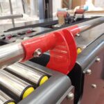 Zbliżenie czerwonego metalowego wspornika przymocowanego do poziomego pręta na taśmie Półautomatyczna zaklejarka Random Tape 50 TBB, pokazująca rolki i części przenośnika w środowisku przemysłowym.