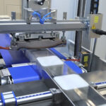 Zbliżenie na Automat pakujący poziomy FLOWPACK SBM750S owijający prostokątne produkty w przezroczystą folię plastikową na przenośniku taśmowym, z niebieskimi i metalowymi elementami z widocznymi etykietami ostrzegawczymi.