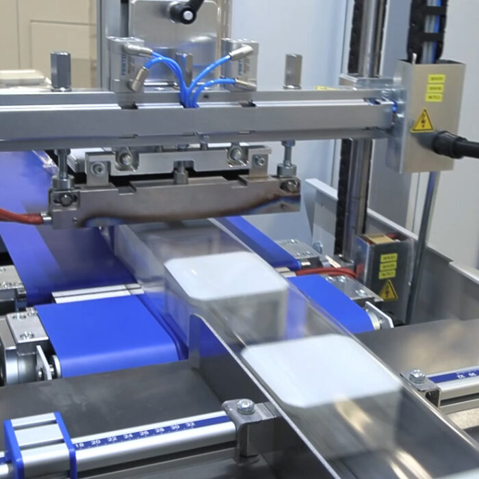 Zbliżenie na Automat pakujący poziomy FLOWPACK SBM750S owijający prostokątne produkty w przezroczystą folię plastikową na przenośniku taśmowym, z niebieskimi i metalowymi elementami z widocznymi etykietami ostrzegawczymi.