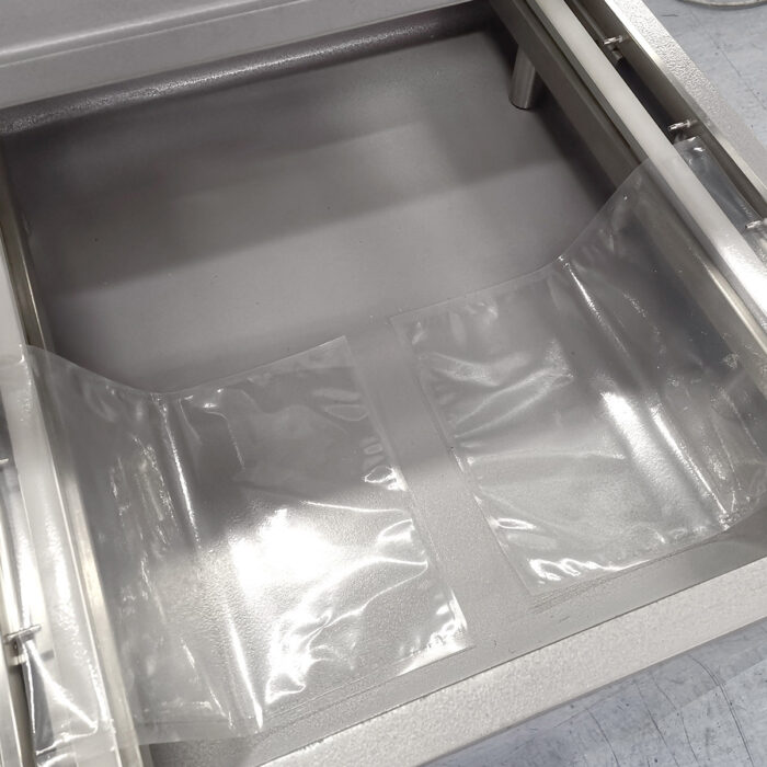 Przezroczyste plastikowe woreczki są umieszczane w komorze ze stali nierdzewnej komorowej pakowarki próżniowej Ultra Vac 600 SA, idealnej do wydajnego zgrzewania próżniowego dzięki gładkiej metalowej powierzchni i widocznym krawędziom maszyny.