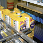 Kilka żółtych opakowań mąki Pão jest owijanych przez Automatyczną zgrzewarkę do zgrzewek FA800AR-150Z, która posiada niebieskie włosie i metalowe ramiona, w otoczeniu przemysłowym.