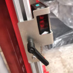 Zbliżenie metalowego uchwytu z czarną dźwignią, zamontowanego na czerwono-srebrnej ramie Półautomatycznej owijarki paletowej PRE-STRETCH Easy Wrap 2000 B, trzymającego urządzenie elektroniczne z widocznym czerwonym czujnikiem.