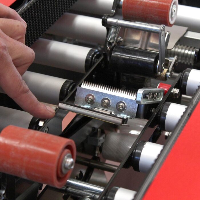Zbliżenie pokazuje palec wskazujący na metalowy mechanizm ostrzy z rolkami, kołami zębatymi i paskami wewnątrz maszyny przemysłowej Półautomatyczna zaklejarka Random Tape 50 SB.