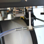 Zbliżenie na półautomatyczną bandownicę stołową Eco Strap 501 z rolkami, szpulą białej taśmy, metalowym prętem i przezroczystą plastikową rurką - pokazujące kluczowe elementy używane w półautomatycznych operacjach pakowania.