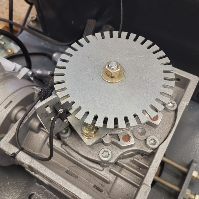 Zbliżenie elementu mechanicznego ze szczelinową okrągłą metalową tarczą, śrubami i czujnikami z czarnymi przewodami, zamontowanego na przekładni Półautomatycznej owijarki palet Slim Wrap 2020B.