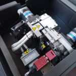 Zbliżenie na półautomatyczną bandownicę stołową Eco Strap 501 C z silnikami elektrycznymi, okablowaniem, etykietami ostrzegawczymi, wentylatorem chłodzącym i metalowymi częściami w ciemnej obudowie. Wysokie napięcie i znaki ostrzegawcze są widoczne.