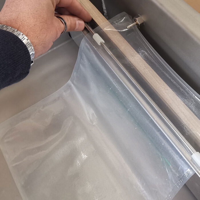 Ręka używa Komorowej pakowarki próżniowej Eco Vac 300S TB do uszczelnienia przezroczystego plastikowego worka za pomocą drewnianego patyczka, pokazanego w zbliżeniu z widocznymi częściami urządzenia i worka.