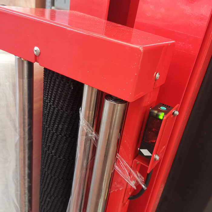 Zbliżenie na owijarkę ramieniową Roto Wrap 1000 AE w kolorze czerwonym, wyposażoną w metalowe pręty, czarny czujnik z zielonym światłem i przezroczystą folię z tworzywa sztucznego owiniętą wokół jednego pręta.