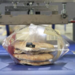 Trzy kawałki płaskiego chleba są układane w przezroczystej plastikowej torbie, zgrzewane przez Automat pakujący poziomy FLOWPACK SBM750S-LR na niebieskim przenośniku taśmowym.