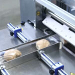 Automatyczna pakowarka flow-pack SR550E pakuje pojedyncze bułki w przezroczystą folię na przenośniku taśmowym w zakładzie przetwórstwa spożywczego.