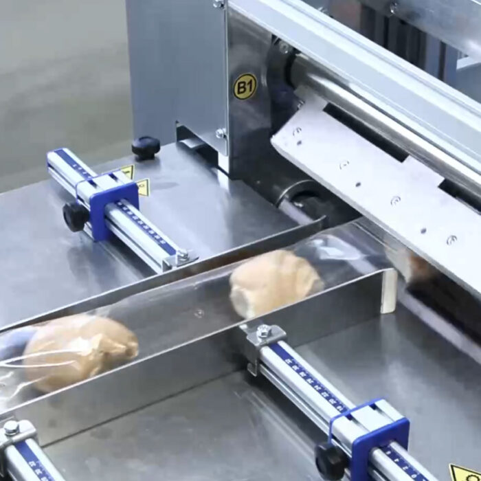 Automatyczna pakowarka flow-pack SR550E pakuje pojedyncze bułki w przezroczystą folię na przenośniku taśmowym w zakładzie przetwórstwa spożywczego.