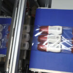 Automatyczna pakowarka flow-pack SR750LR owija plastikowe butelki z czerwonymi nakrętkami w przezroczysty plastik na przenośniku taśmowym w zakładzie pakowania.