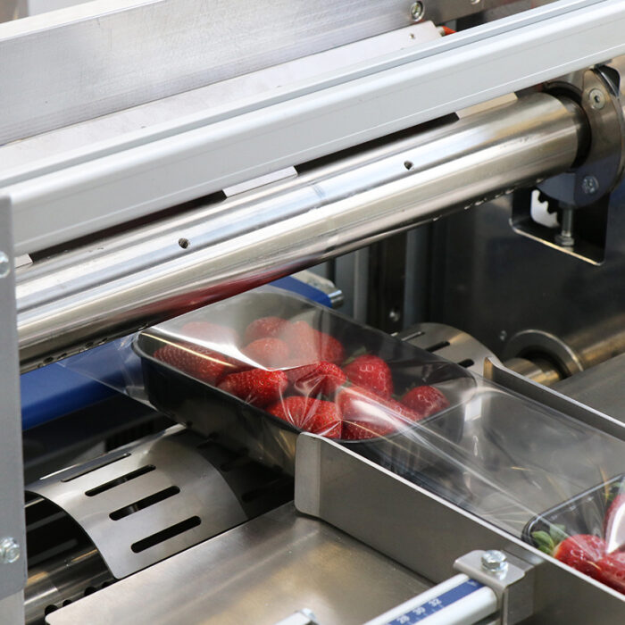 Zbliżenie na Automatyczną pakowarkę flow-pack SR550 owijającą świeże truskawki w przezroczysty plastikowy pojemnik, z folią częściowo przykrywającą owoce, gdy poruszają się wzdłuż przenośnika.