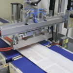 Zbliżenie na Automatyczną pakowarkę flow-pack SR550LR owijającą przedmioty w przezroczystą folię plastikową na niebieskim przenośniku taśmowym w fabryce, z widocznymi metalowymi częściami, kablami i etykietami ostrzegawczymi.