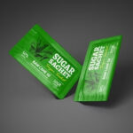 Dwa zielone opakowania SUGAR SACHET (50% RAW CANE, 10GR, obrazy trzciny cukrowej) spoczywają na ciemnoszarej powierzchni - każde wykonane przez Pionowa maszyna pakująca Mini Vert 240 DP.