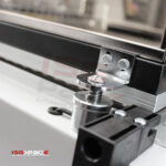 Zbliżenie metalowego mechanizmu zatrzaskowego ze sprężynową śrubą i wspornikiem montażowym na obramowanym szklanym panelu, część Zgrzewarki kloszowej do folii Easy Sealer 85. Logo ISG PACK jest widoczne w lewym dolnym rogu.