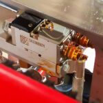 Zbliżenie metalowego elementu z Automatycznej zaklejarki Matic Tape 50 CF HM, oznaczonego jako SA23MB5671, z kodem QR, złotymi złączami i przewodami przymocowanymi do metalowej ramki.