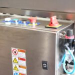 Zbliżenie panelu sterowania ze stali nierdzewnej w Automatycznej zaklejarce Auto Tape 50 CF SS, pokazujące kolorowe przyciski, wyłącznik awaryjny, etykiety ostrzegawcze, dołączone rurki, blokadę i regulator ciśnienia po prawej stronie.