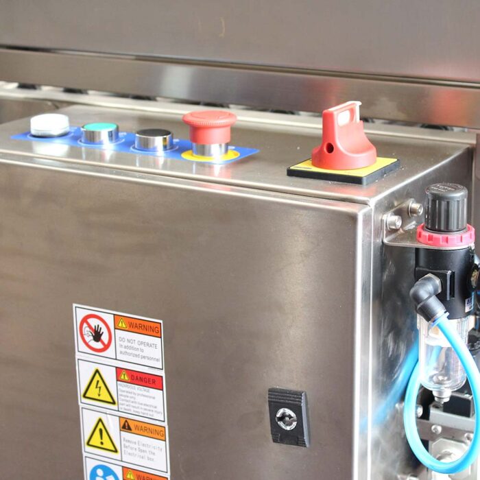Zbliżenie panelu sterowania ze stali nierdzewnej w Automatycznej zaklejarce Auto Tape 50 CF SS, pokazujące kolorowe przyciski, wyłącznik awaryjny, etykiety ostrzegawcze, dołączone rurki, blokadę i regulator ciśnienia po prawej stronie.