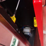 Zbliżenie czerwonej metalowej osłony maszyny z dwoma żółtymi czujnikami bezpieczeństwa SICK monitorującymi otwieranie i zamykanie osłony na półautomatycznej bandownicy do palet Pallet Strap T150 dla bezpieczeństwa.