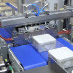 W fabryce Automat pakujący poziomy FLOWPACK SI550S owija plastikowy pojemnik przezroczystą folią na niebieskim przenośniku taśmowym.