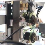 Zbliżenie metalowych prętów, przewodów, złączy i kabli na szpuli z Pionowa maszyna pakująca Mini Vert 240 PP, pokazujące różne metalowe i mechaniczne elementy w jej montażu.