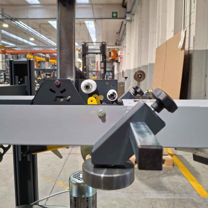 Zbliżenie Półautomatyczna zaklejarka kartonów SIAT S8 z metalowymi częściami i pokrętłami sterującymi w magazynie, otoczona półkami, sprzętem i wózkiem widłowym w jasnym oświetleniu w pomieszczeniu z wysokim sufitem.