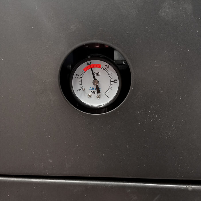 Zbliżenie okrągłego manometru na ciemnej metalowej powierzchni pokazuje odczyty MPa i ATM. Igła wskazuje czerwoną strefę w pobliżu zera, wskazując niskie ciśnienie - idealne do monitorowania Owijarka do paczek Box Wrap 800 B.